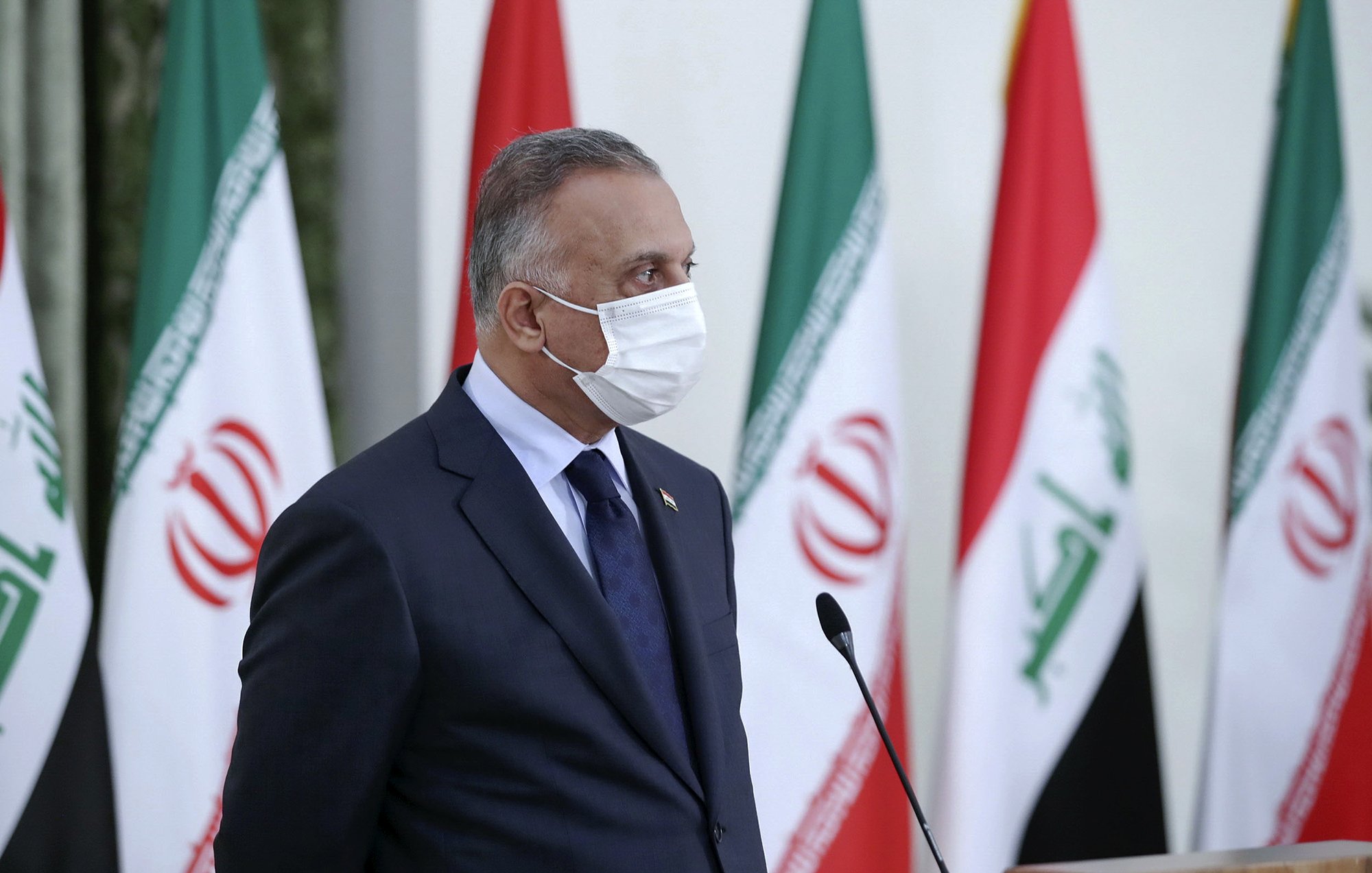 The image depicts a person wearing a mask, standing in profile. He is dressed in a dark suit with a tie and appears to be engaged in an official event. In the background, multiple flags are visible, including those of Iran and Iraq, suggesting a diplomatic or governmental setting. The person appears serious and focused, possibly addressing an audience or participating in a press conference.