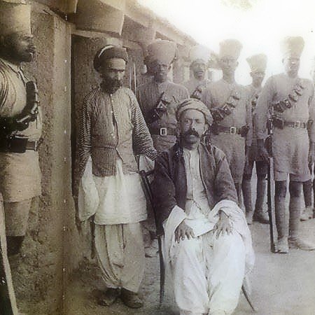 الصورة تظهر مجموعة من الرجال يرتدون زيًا عسكريًا تقليديًا، يقفون في صفوف. في المقدمة، يجلس رجل على كرسي، يبدو أنه شخصية ذات مكانة أو أهمية. يرتدي الرجل ملابس تقليدية مختلفة عن الآخرين، مما يدل على اختلاف اجتماعي أو ثقافي. الخلفية تبدو بسيطة، وتضيف إلى الطابع التاريخي للصورة. الأجواء تتسم بالجدية، ويبدو أن الصورة قد التُقطت في فترة زمنية سابقة.