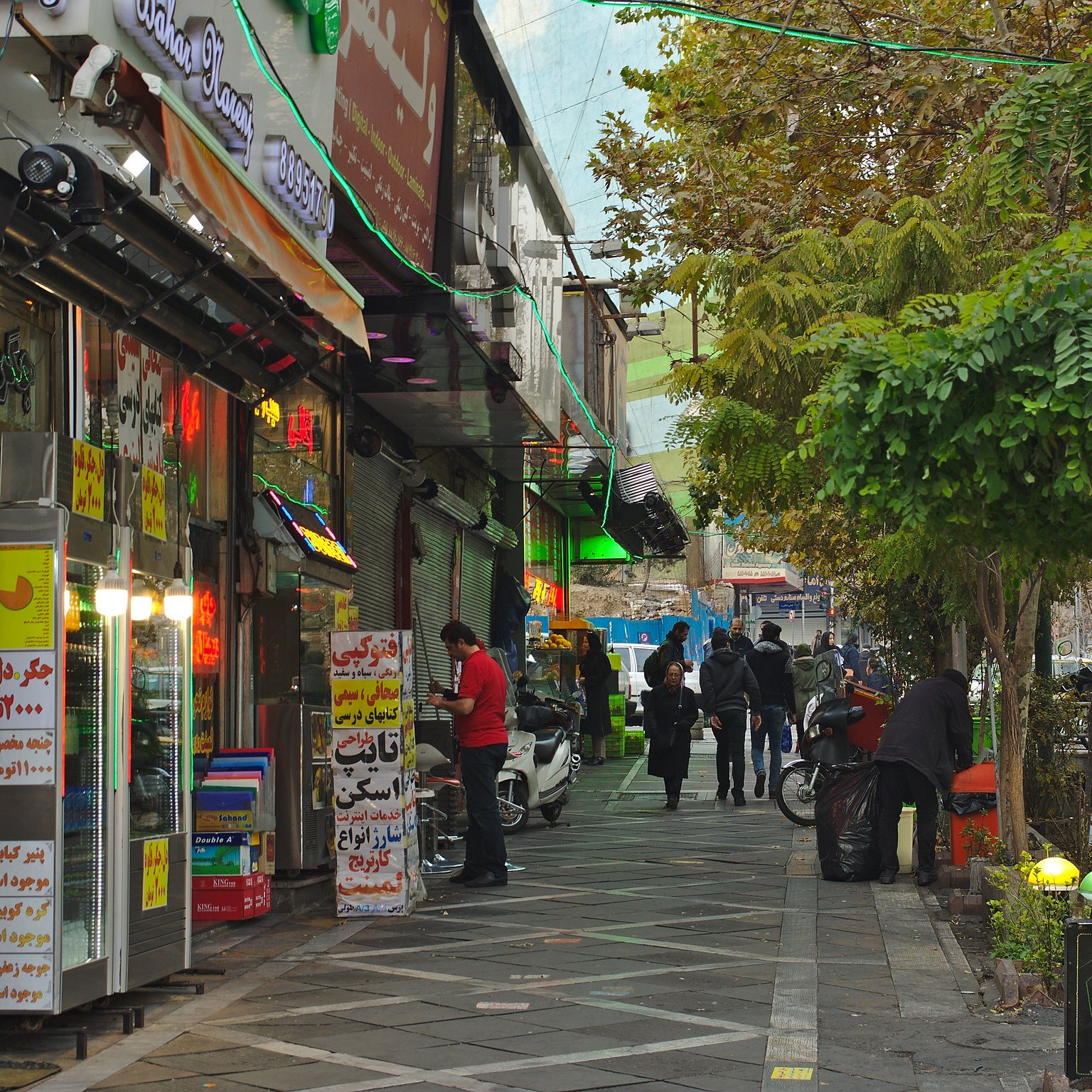 Une rue animée bordée de magasins colorés et de personnes marchant sous des arbres.