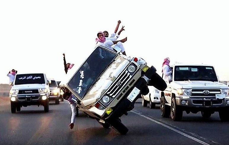 L'image montre plusieurs véhicules tout-terrain sur une route. Un des véhicules est en train de faire un équilibrage sur deux roues, avec des personnes à l'intérieur et à l'extérieur qui semblent célébrer et s'amuser. Les passagers portent des vêtements traditionnels, et l'ambiance générale semble festive. En arrière-plan, d'autres véhicules sont visibles, créant une scène dynamique et animée.