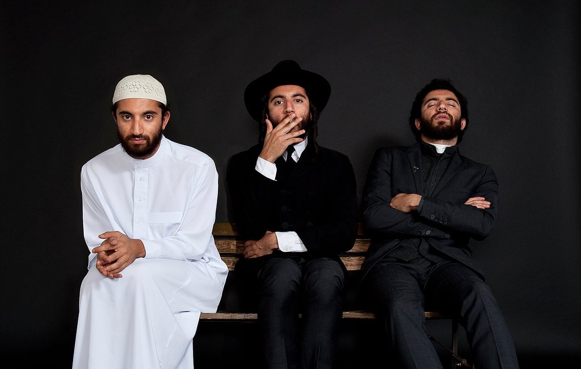 L'image montre trois hommes assis sur un banc, chacun portant un style vestimentaire distinct. À gauche, un homme en blanc, portant une tenue traditionnelle et un couvre-chef. Au centre, un homme habillé de manière formelle, avec un chapeau noir et une tenue sombre. À droite, un homme également en costume, avec une expression pensive. Le fond est noir, ce qui met en valeur les vêtements et les postures des sujets.