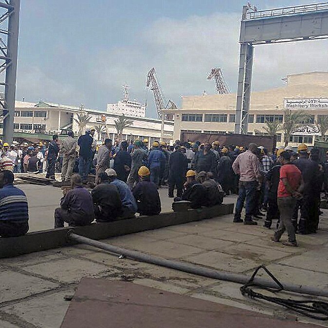 L'image montre un groupe de nombreux travailleurs rassemblés dans un espace extérieur, probablement dans un port ou un chantier naval. Les personnes sont vêtues de tenues de travail et portent des casques de sécurité jaunes. Certains d'entre eux sont assis sur le sol, tandis que d'autres sont debout, discutant ou attendant. L'environnement semble industriel, avec des structures métalliques et des équipements visibles en arrière-plan. Le ciel est partiellement nuageux.