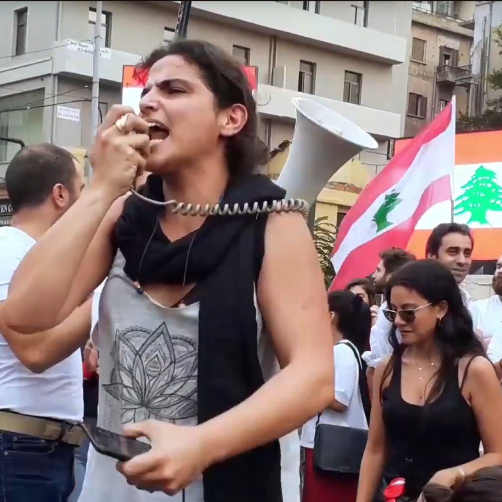 Foule en manifestation, femme au mégaphone, drapeaux libanais en arrière-plan.