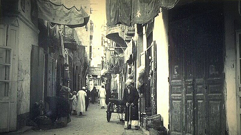 L'image montre une ruelle étroite d'une ville ancienne, probablement dans un pays arabe. Les murs des bâtiments sont ornés de tentures et de draps qui ajoutent une ambiance colorée. On peut voir des habitants, dont certains portent des vêtements traditionnels, déambulant dans la rue. Un homme semble se tenir près d'une charrette, tandis que d'autres personnes se déplacent en arrière-plan. Les portes en bois, typiques, ajoutent un charme ancien à la scène, qui évoque une atmosphère de vie quotidienne dans un vieux quartier.