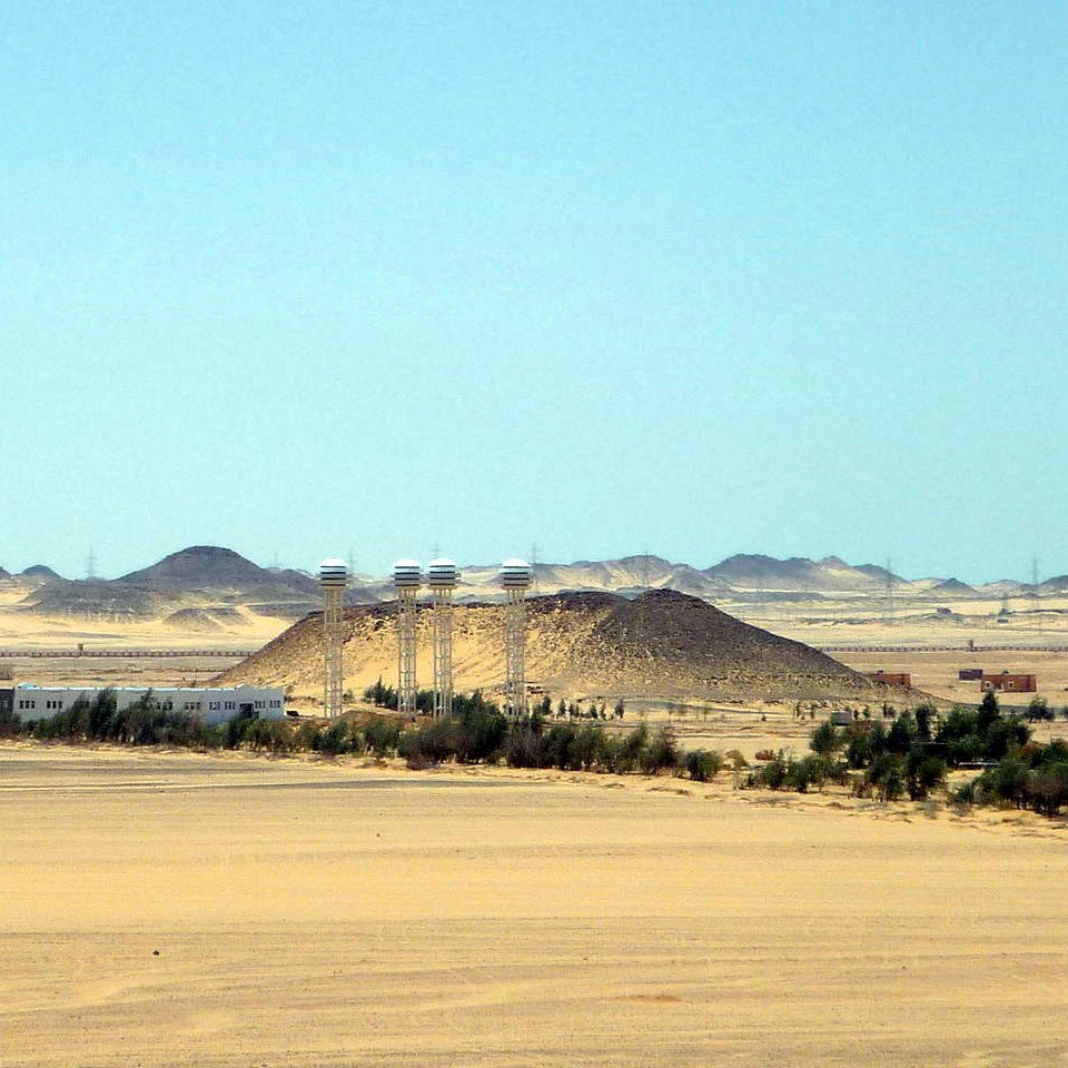 L'image montre un paysage désertique vaste et aride, avec des dunes de sable et des collines. Au centre, on peut distinguer quelques bâtiments qui semblent être des installations humaines, entourés de végétation telle que des arbres ou des buissons. À l'arrière-plan, des collines se découpent contre le ciel, tandis qu'une lumière vive souligne l'immensité du paysage. L'atmosphère semble calme et isolée, typique des régions désertiques.