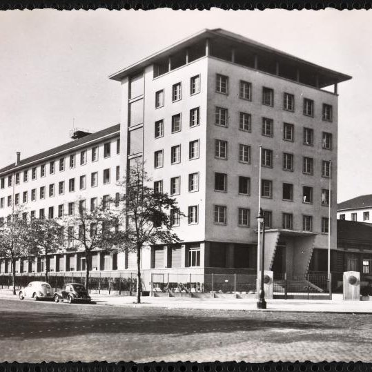 L'image représente un bâtiment architectural de plusieurs étages, probablement de style moderne. Il est photographié sous une lumière naturelle, mettant en avant sa façade aux lignes géométriques. Le bâtiment semble être entouré d'arbres et de quelques véhicules anciens, ce qui donne une ambiance d'époque. On peut également apercevoir des éléments de la rue, comme des lampadaires et éventuellement d'autres bâtiments en arrière-plan. L'ensemble de l'image présente une esthétique monochrome, accentuant le caractère historique de la scène.