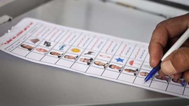 L'image montre une main tenant un stylo en train de remplir un bulletin de vote. Le bulletin affiche plusieurs colonnes, chacune contenant des photos de différentes personnes, accompagnées de symboles et de cases à cocher. Cela semble être un contexte électoral, où une personne vote pour des candidats. Le bulletin est posé sur une surface grise.