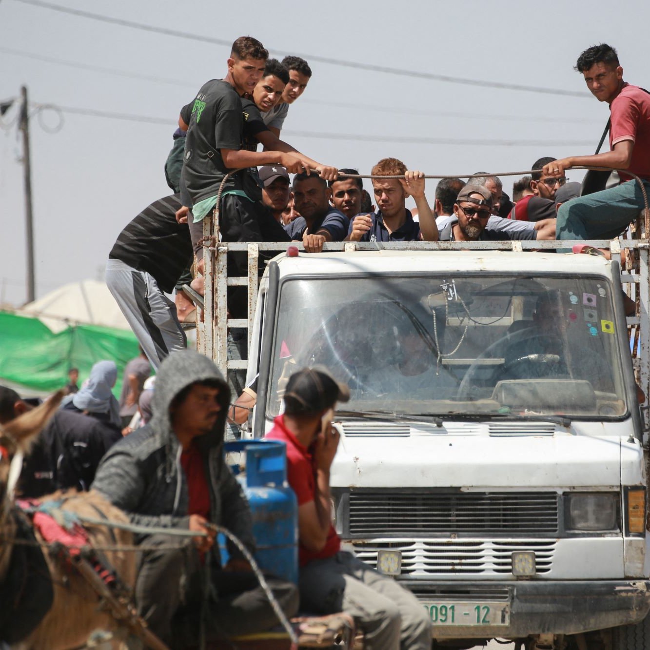 L'image montre un groupe de personnes assises sur le toit d'un camion, tandis que d'autres se trouvent à proximité, y compris des personnes à pied et une charrette tirée par un cheval. L'environnement semble être un endroit animé, avec des tentes et des structures visibles en arrière-plan. On peut voir des fils électriques et des montagnes de déchets, ce qui suggère une atmosphère informelle ou de camp. La scène dégage une impression de communauté et de vie quotidienne dans un contexte peut-être difficile.