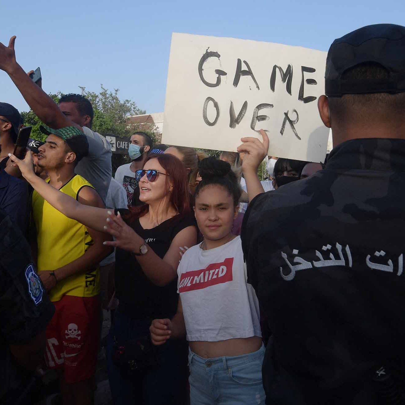 تظهر الصورة تجمعًا لعدد من الأشخاص في مُظاهرة، حيث يبدو أن هناك تبادلًا للآراء بين المحتجين والقوات الأمنية. بعض المتظاهرين يحملون لافتات، بينما يبدو آخرون يتفاعلون بشكل نشط مع الوضع. هناك تنوع في ملابس المشاركين، مما يوحي بتعدد الأعمار والجنسيات. تعكس الصورة أجواء من الحماس والاحتجاج، حيث يُظهر المحتجون رغبتهم في التعبير عن وجهات نظرهم.