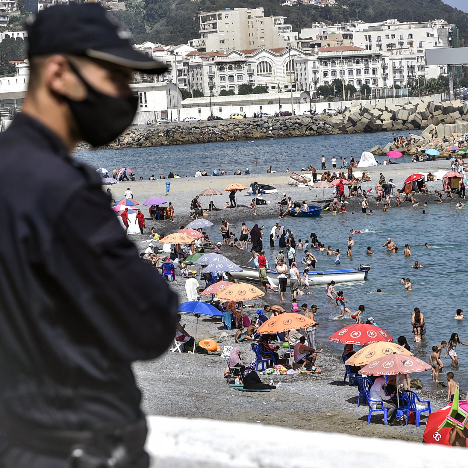 تظهر الصورة شاطئاً مزدحماً حيث يتواجد العديد من الأشخاص الذين يستمتعون بالسباحة والاسترخاء تحت الشمس. يوجد في الصورة مجموعة من المظلات الملونة التي توفر الظل للزوار. في المقدمة، هناك رجل أمن يرتدي كمامة وبدلة رسمية، يراقب الأوضاع. يظهر في الخلفية مجموعة من المباني والجبال، مما يضفي طابعاً جميلاً على المشهد.