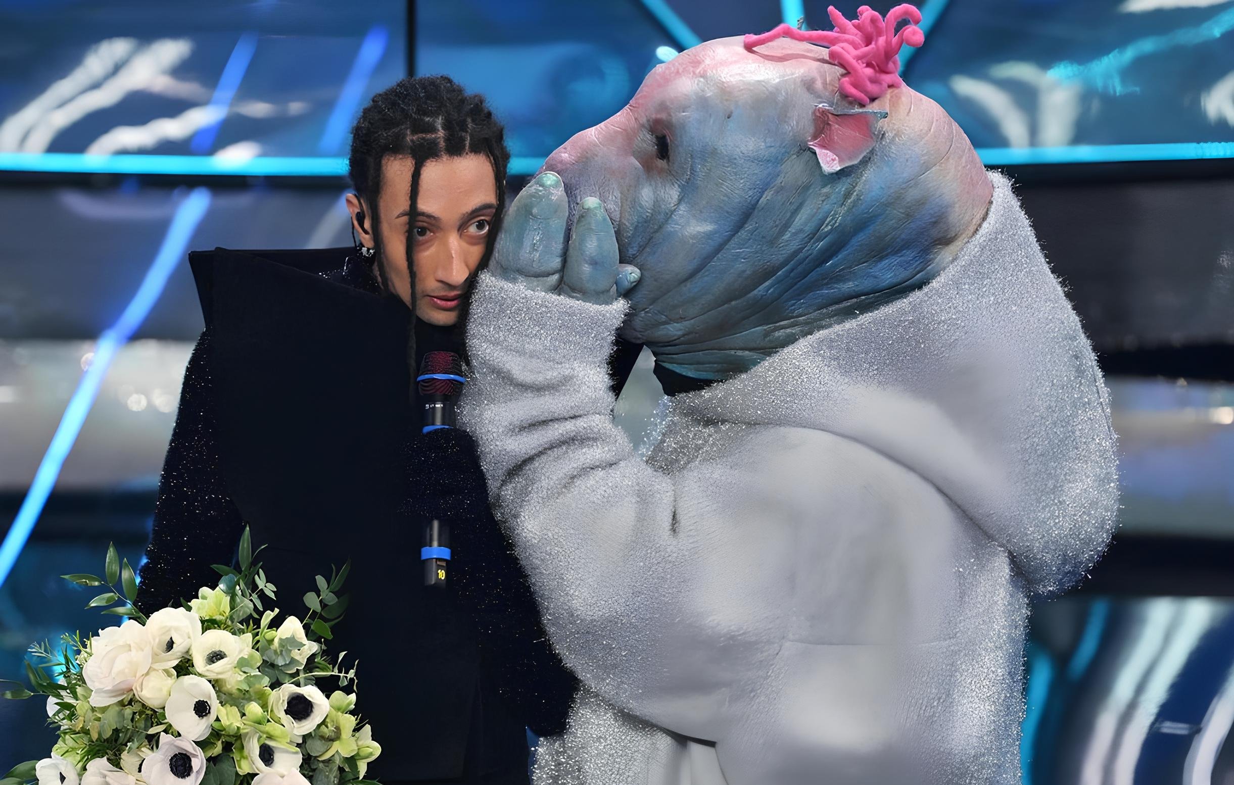 L'immagine mostra due figure in un ambiente scenico. Una persona, vestita con abiti scuri e con capelli lunghi e intrecciati, tiene un microfono e un mazzetto di fiori. Accanto a lei c'è un personaggio mascherato, che indossa un costume piumato e una maschera con tratti insoliti. Il personaggio sembra sussurrare all'orecchio della persona, creando un'atmosfera di intimità o mistero. Sullo sfondo si possono vedere luci blu e decorazioni moderne, tipiche di uno spettacolo televisivo.