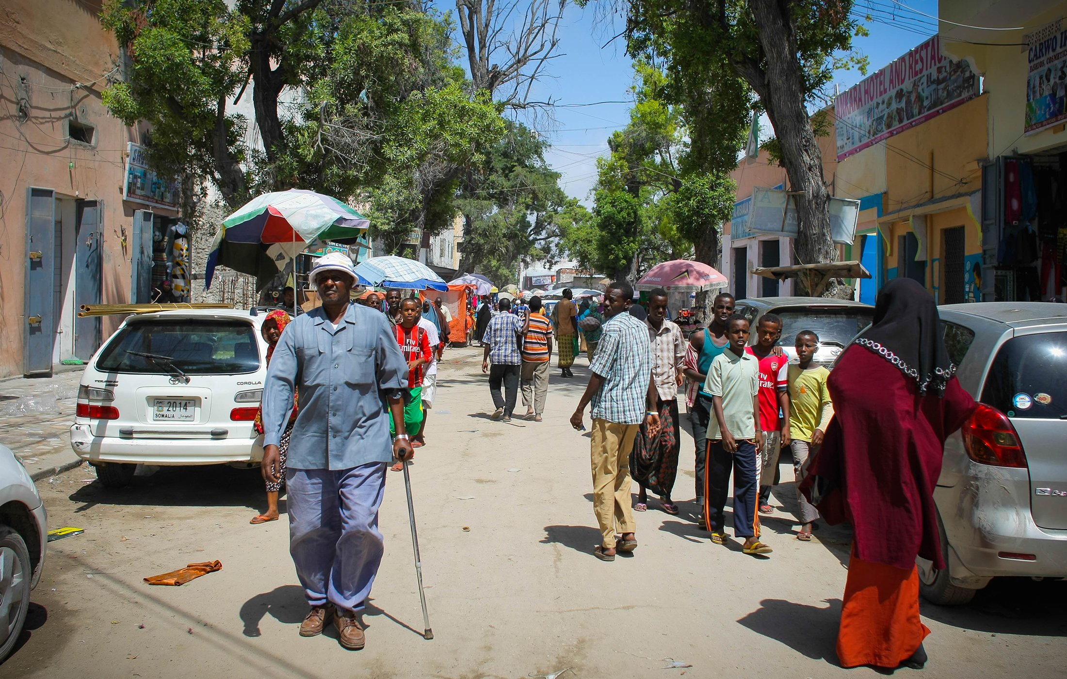 L'image montre une rue animée avec des groupes de personnes marchant. On peut voir un homme âgé utilisant une canne, habillé en bleu, qui semble avancer lentement. Plusieurs personnes sont dispersées le long de la rue, certaines se trouvent près de voitures garées. Des ombrelles colorées sont visibles, probablement pour se protéger du soleil. Les bâtiments en arrière-plan semblent être de petits commerces ou habitations. L'ambiance générale dénote une vie quotidienne dynamique dans un environnement urbain.