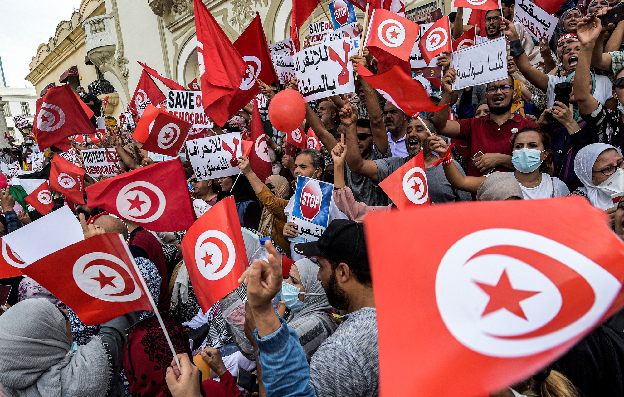 L'image montre une foule manifestante en Tunisie, brandissant des drapeaux tunisiens et des pancartes. Les participants expriment des messages en faveur de la démocratie, avec des slogans tels que "Sauvez la démocratie" et "Stop". On peut voir des gens de différents âges et genres, certains portant des masques. L'ambiance semble énergique et engagée, avec un fort esprit de revendication collective.