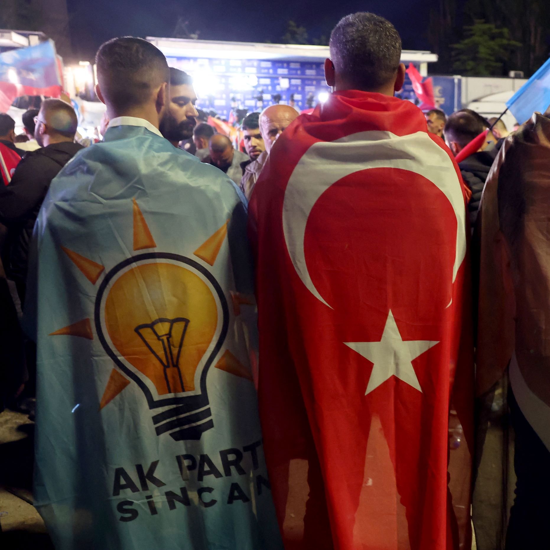 در این تصویر، چند نفر دیده می‌شوند که از دو نوع لباس مخصوص استفاده کرده‌اند. یکی از آنها پرچم ترکیه را به دوش انداخته و دیگری با تصویری از رجب طیب اردوغان، رئیس‌جمهور ترکیه، پوشیده شده است. این افراد در یک تجمع سیاسی حضور دارند، به نظر می‌رسد که از طرفداران حزب عدالت و توسعه (AKP) هستند و فضای شاد و ملی‌گرایانه‌ای در اطرافشان حاکم است. تعدادی پرچم و نمادهای دیگری هم در پس‌زمینه دیده می‌شود.