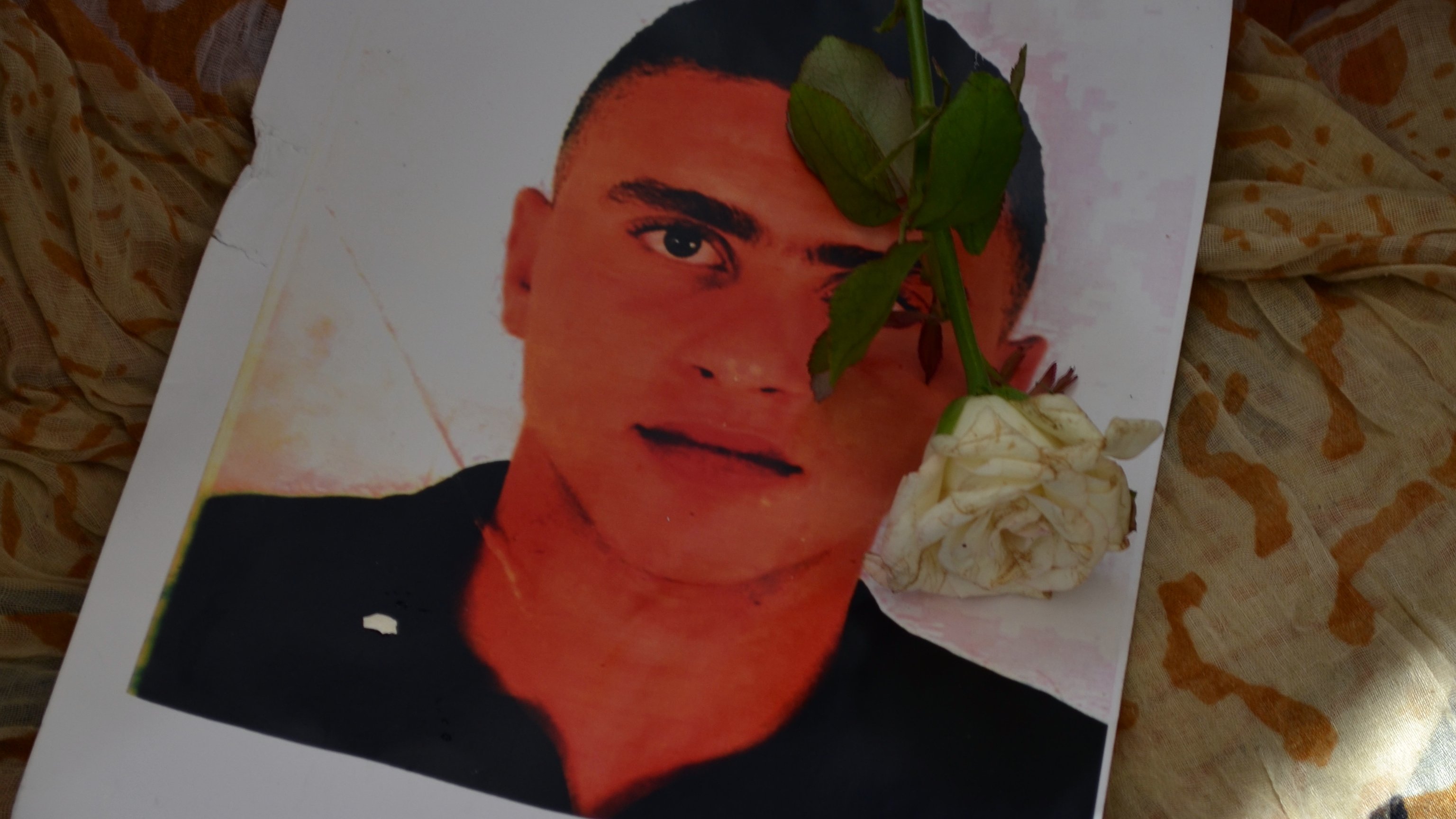 L'image montre un portrait d'un jeune homme, placé sur un tissu aux motifs colorés. Une rose blanche est délicatement posée sur le portrait, ajoutant une touche de sérénité et de respect. Les couleurs et les textures du tissu environnant contrastent avec l'image du jeune homme.