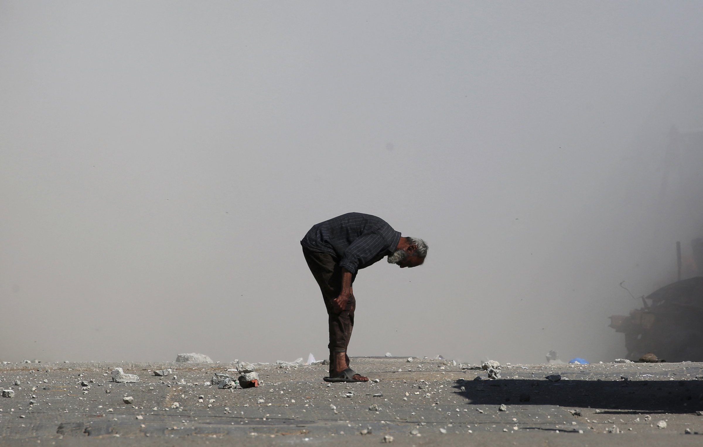 Un uomo anziano si piega in avanti su una strada polverosa, circondato da detriti.