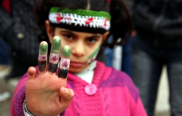 L'image montre une jeune fille qui lève la main en avant, exhibant ses doigts teintés de peinture. Elle porte un pull en laine rose et un bandeau coloré orné de motifs. Son visage est marqué par des traces de peinture, notamment sur les joues. L'expression de son visage semble déterminée, et elle attire l'attention sur son geste. En arrière-plan, on peut apercevoir des silhouettes floues de personnes, suggérant un rassemblement ou une manifestation.