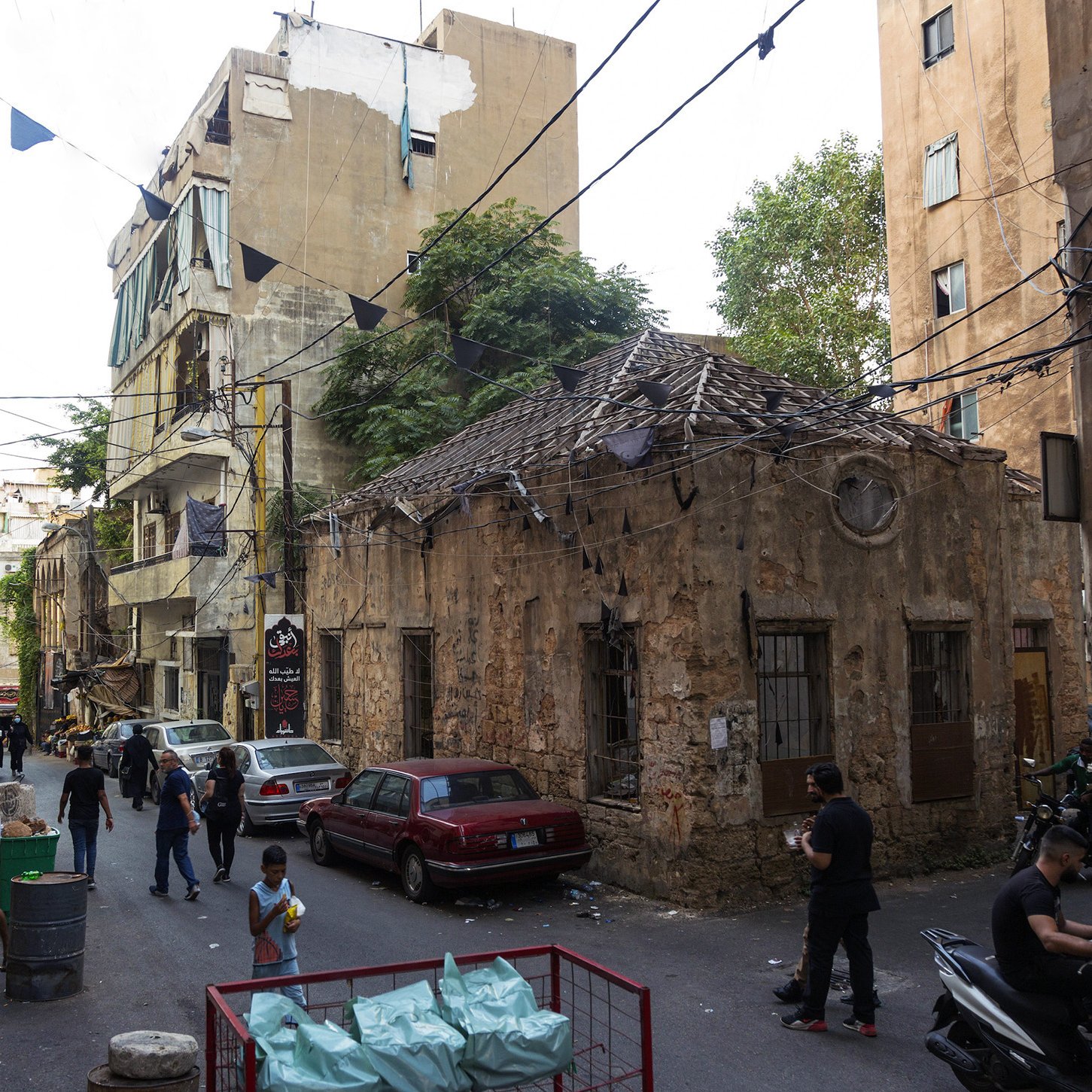 L'image montre une ruelle étroite dans une ville, probablement au Liban. On peut y voir des bâtiments anciens avec des murs en pierre usée par le temps. Certains bâtiments semblent en mauvais état, tandis que d'autres sont légèrement rénovés. La rue est animée avec des personnes qui marchent, certaines d'entre elles se tenant près de véhicules stationnés. Des banderoles pendant au-dessus de la rue ajoutent une touche de couleur. En général, l'atmosphère semble à la fois vivante et marquée par l'histoire.