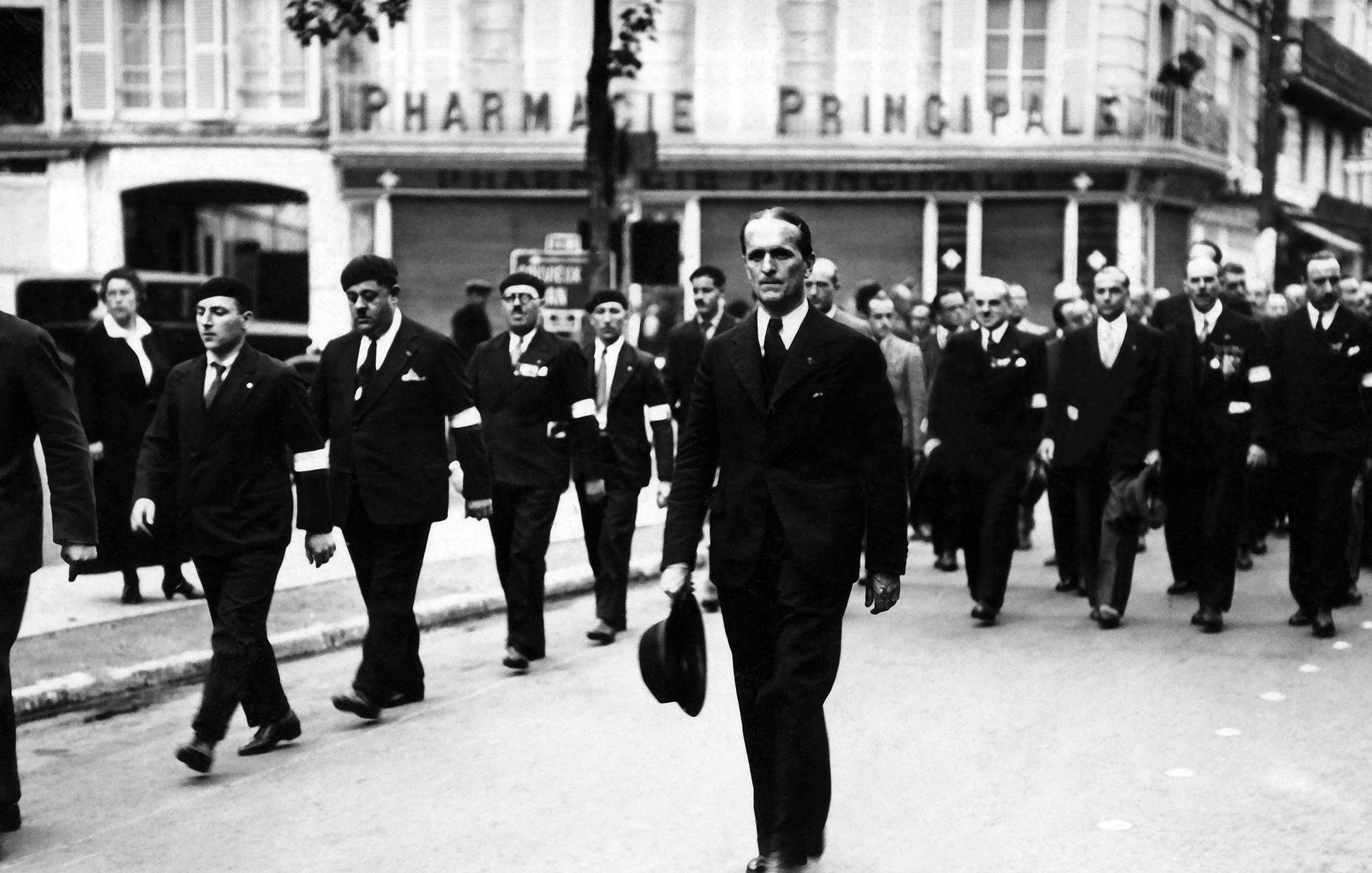 L'image montre un groupe de personnes défilant dans une rue, probablement lors d'un événement formel ou d'une commémoration. Au premier plan, un homme en costume noir porte un chapeau et marche avec assurance. Les autres participants portent aussi des costumes et certains semblent avoir des insignes ou des decorations. En arrière-plan, on peut voir un bâtiment avec l'enseigne "PHARMACIE PRINCIPALE". L'atmosphère générale de la scène semble sérieuse et respectueuse.