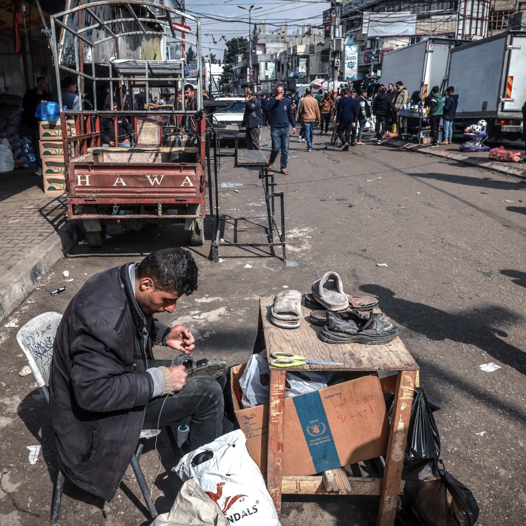 L'image montre une scène de rue animée. Au premier plan, un homme est assis sur une chaise, concentré sur sa tâche. Il répare des chaussures, utilisant des outils comme des ciseaux et des filaments. Devant lui, une table supporte des chaussures à réparer et un sac. À l'arrière-plan, on peut voir des personnes marchant le long d'une rue entourée de camions et de petites échoppes. L'atmosphère semble vivante, avec des éléments de la vie quotidienne qui se déroulent autour de lui.