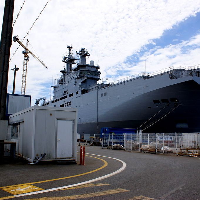 L'image montre un grand navire de guerre, probablement un porte-hélicoptères ou un navire amphibie, amarré dans un port. Le navire est de couleur grise et possède des structures sur le pont, suggérant qu'il est conçu pour des opérations militaires. En arrière-plan, on peut voir des grues et d'autres installations portuaires, tandis qu'à l'avant, il y a un petit bâtiment et des barrières de sécurité. Le ciel est partiellement nuageux, ajoutant une ambiance de chantier naval.