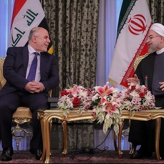 تظهر الصورة لقاءً بين شخصين يجلسان على طاولة مزينة بزهور. العلم العراقي وعلم إيران خلفهم، مما يشير إلى أن الاجتماع يعقد بين ممثلين عن البلدين. يبدو أن كلا الشخصين يستمتعان بالنقاش ولديهما تعابير وجه إيجابية. الديكور في الخلفية أنيق ومزخرف، مما يعكس طابعًا رسميًا لهذا اللقاء.