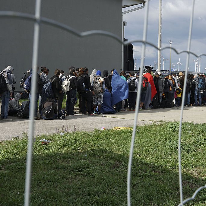 L'image montre une file de personnes faisant la queue près d'un bâtiment, probablement en attendant de recevoir de l'aide ou des services. Les personnes sont vêtues de diverses manières, avec certains portant des couvertures. On peut voir une clôture en fil de fer qui sépare le groupe du reste de l'environnement. En arrière-plan, on aperçoit des installations modernes et des éoliennes, suggérant une zone urbaine ou industrielle. Le ciel est nuageux, ce qui pourrait indiquer une atmosphère morose.