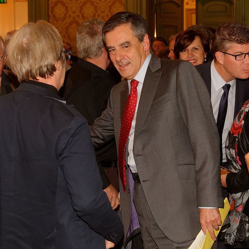 L'image montre un groupe de personnes réunies dans une salle, probablement lors d'un événement. Au centre, un homme en costume, avec une cravate à pois rouges, semble engager une conversation avec quelqu'un en lui serrant la main. D'autres personnes, en arrière-plan, observent ou participent à la discussion. L'ambiance semble conviviale, avec des sourires et des échanges entre les participants. Les murs sont décorés et ajoutent une touche d'élégance à la scène.