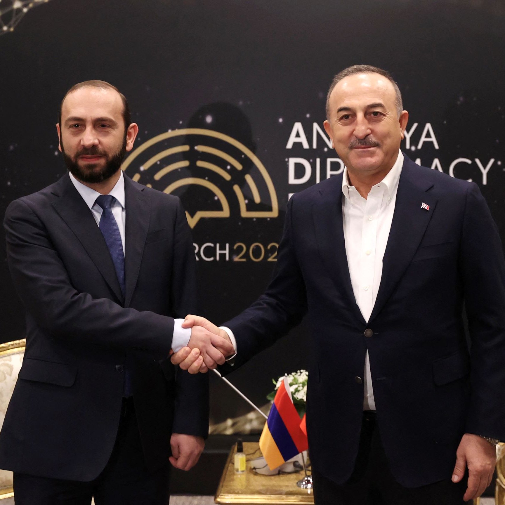 تظهر الصورة شخصين يتصافحان في مناسبة دبلوماسية. يقف الشخصان أمام خلفية تحمل عبارة "أنقرة دبلوماسية" وتاريخ مارس 2022. يرتدي أحدهما بدلة رسمية بينما يبدو الآخر أيضًا في زي رسمي. المكان يبدو أنيقًا، ويوجد خلفهما تصميم مميز يبرز الطابع الرسمي للاجتماع.