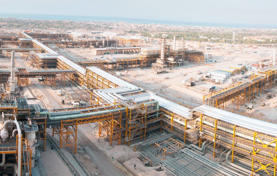 L'image montre un vaste complexe industriel, probablement lié à l'extraction ou au traitement des ressources naturelles. On peut voir de nombreuses structures métalliques, des tubes et des pipelines s'étendant sur une grande surface. L'environnement semble désertique ou aride, et au loin, on aperçoit la mer. La disposition des installations indique un environnement de travail organisé, avec des infrastructures complexes qui témoignent d'une activité industrielle importante.