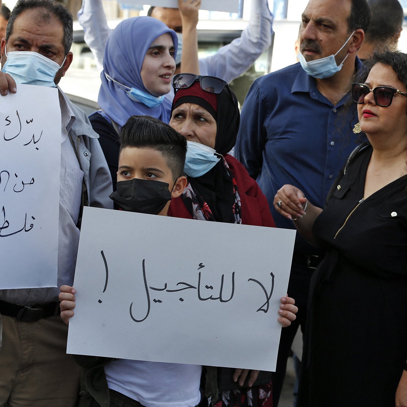 تظهر في الصورة مجموعة من الناس في مظاهرة. يرفع بعضهم لافتات، بينما يرتدي الآخرون كمامات. في المقدمة، يوجد طفل صغير يحمل لافتة مكتوب عليها "لا للتأجيل!"، وتحيط به مجموعة من البالغين، بينهم امرأة ترتدي فستانًا أسود وبعض الرجال بملابس تقليدية. تعكس الصورة جوًا من الاحتجاج والتعبير عن الرأي.