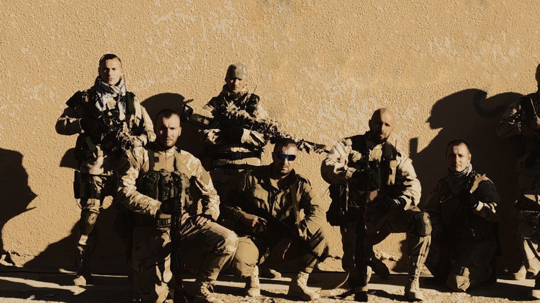 L'image montre un groupe de soldats en tenue militaire. Ils sont positionnés à la fois debout et accroupis, avec des armes à feu en main. L'arrière-plan est une surface murale de couleur beige, ce qui suggère un environnement désertique. Les soldats portent des uniformes camouflés et semblent prêts pour une mission. L'éclairage est plutôt clair, mettant en valeur leurs expressions et leurs équipements.