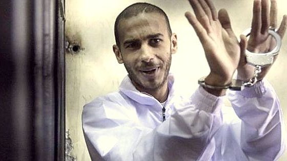 L'image montre un homme au regard confiant, portant une combinaison blanche. Ses mains sont menottées, et il semble faire un geste de la main, peut-être en souriant ou en se moquant de la situation. Le fond de l'image est flou, ce qui met l'accent sur l'homme. L'atmosphère générale donne une impression d'intensité, et l'homme semble détaché ou à l'aise malgré les menottes.