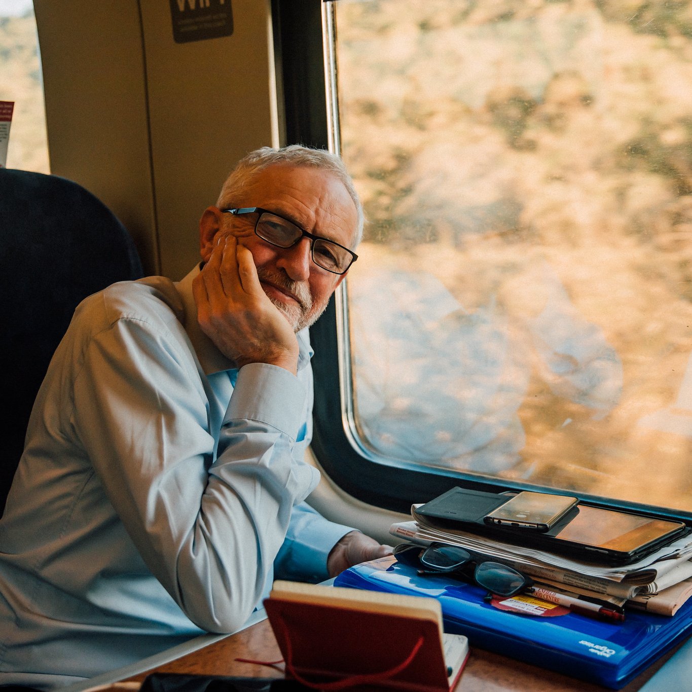 L'image montre un homme assis à côté d'une fenêtre de train. Il porte une chemise claire et a des lunettes. Son visage exprime une certaine réflexion ou contemplation alors qu'il repose sa tête sur sa main. Sur la table devant lui, on peut voir plusieurs objets comme des téléphones, des lunettes de soleil et des papiers, donnant l'impression qu'il a passé du temps à travailler ou à organiser ses affaires. À l'extérieur de la fenêtre, on aperçoit un paysage flou, suggérant que le train est en mouvement.