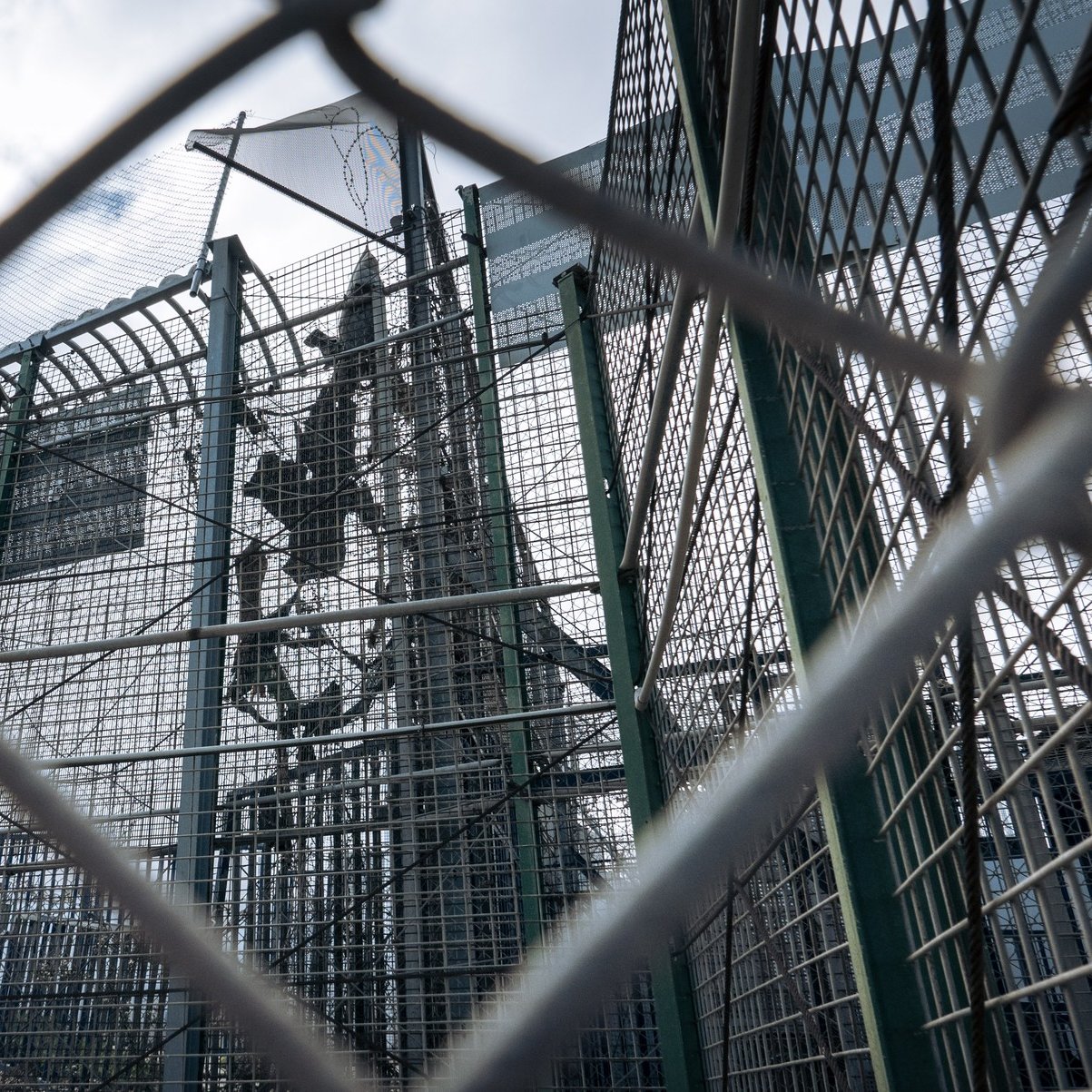 L'image montre une structure en métal, probablement une clôture ou un mur de sécurité, entourée de grillages. On peut apercevoir des barres et une superficie qui semble être une installation sécurisée. Le ciel est nuageux, ce qui donne une atmosphère un peu sombre à la scène. Cette image évoque un environnement restreint, probablement lié à la sécurité ou à la protection.