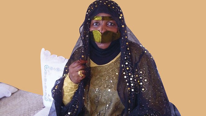 الصورة تظهر امرأة ترتدي زيًا تقليديًا مميزًا، والذي يتضمن عباءة سوداء تغطي جسدها وزينتها بألوان ذهبية أو لامعة. وجهها مغطى جزئيًا بحجاب، مما يبرز فقط عينيها. المرأة تجلس في بيئة منزلية بسيطة، حيث توجد بعض الأواني والزينة حولها. تعكس الصورة تراثًا ثقافيًا غنيًا وتفاصيل تقليدية لعادات معينة.