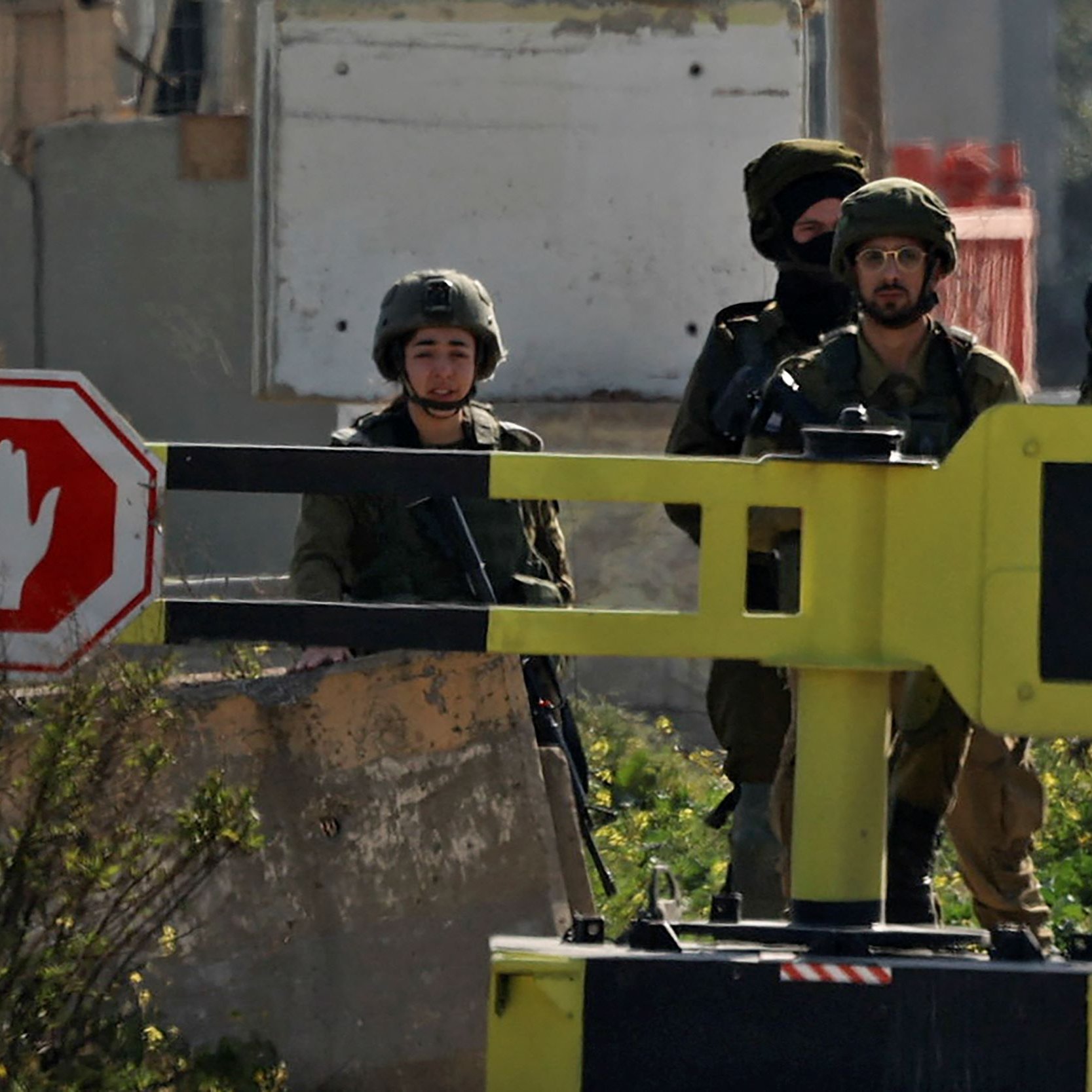 در این تصویر، گروهی از نظامیان در یک خیابان یا منطقه‌ای مستقر هستند. در جلوی آن‌ها، یک علامت ترافیکی به شکل علامت «ایست» قرار دارد. نظامیان در حالت آماده‌باش به نظر می‌رسند و تعدادی از آن‌ها سلاح در دست دارند. محیط اطراف شامل دیوارها و سازه‌های مختلف است که نشان‌دهنده یک منطقه نظامی یا امنیتی می‌باشد. فضای کلی تصویر تنش‌زا و جدی به نظر می‌رسد.