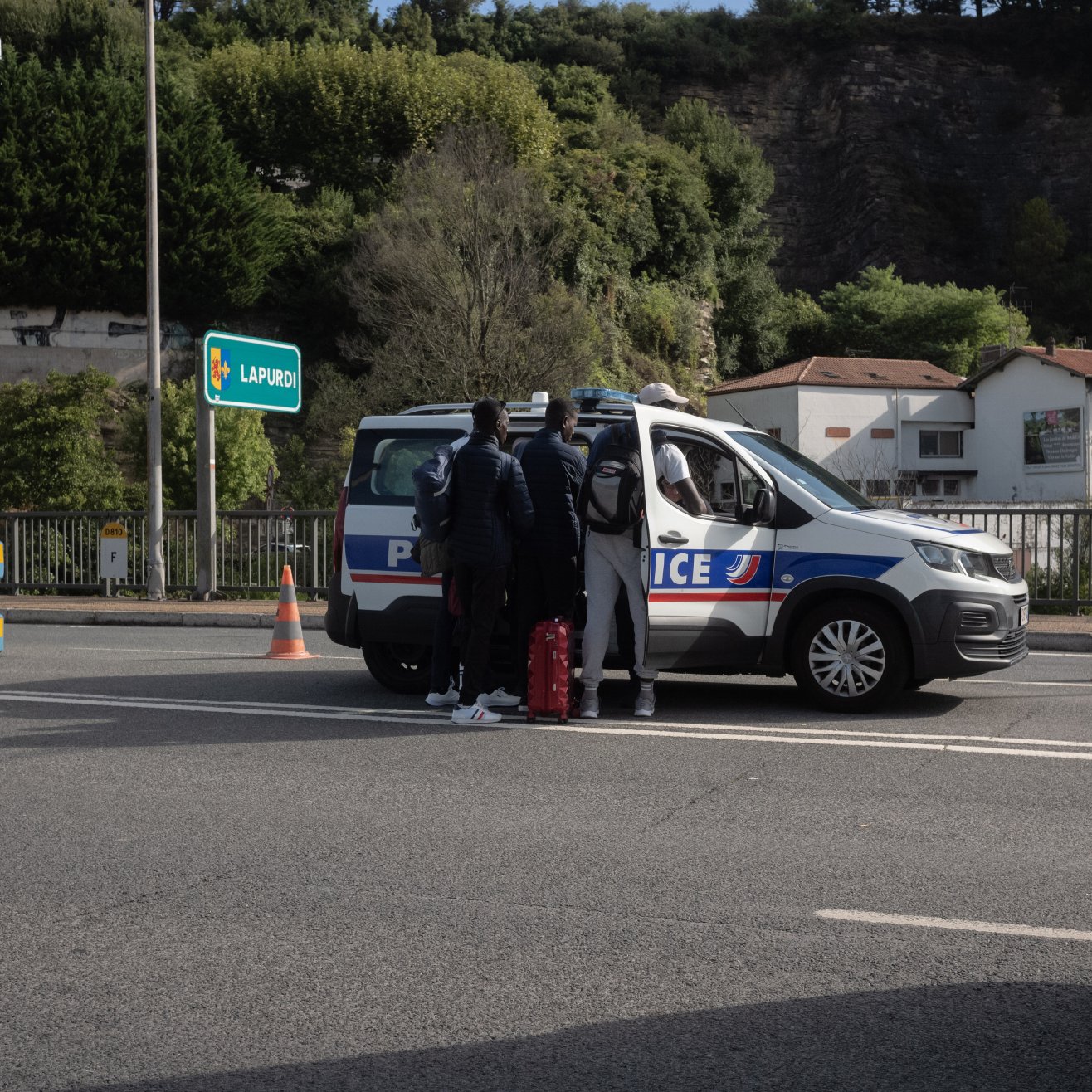 L'image montre une scène sur une route, où un véhicule de police est stationné. Des personnes se tiennent près de la voiture de police, probablement en train de discuter avec les agents. En arrière-plan, on peut voir quelques bâtiments et de la végétation. Des cônes de signalisation sont visibles sur la voie, indiquant peut-être une opération de contrôle ou de sécurité.