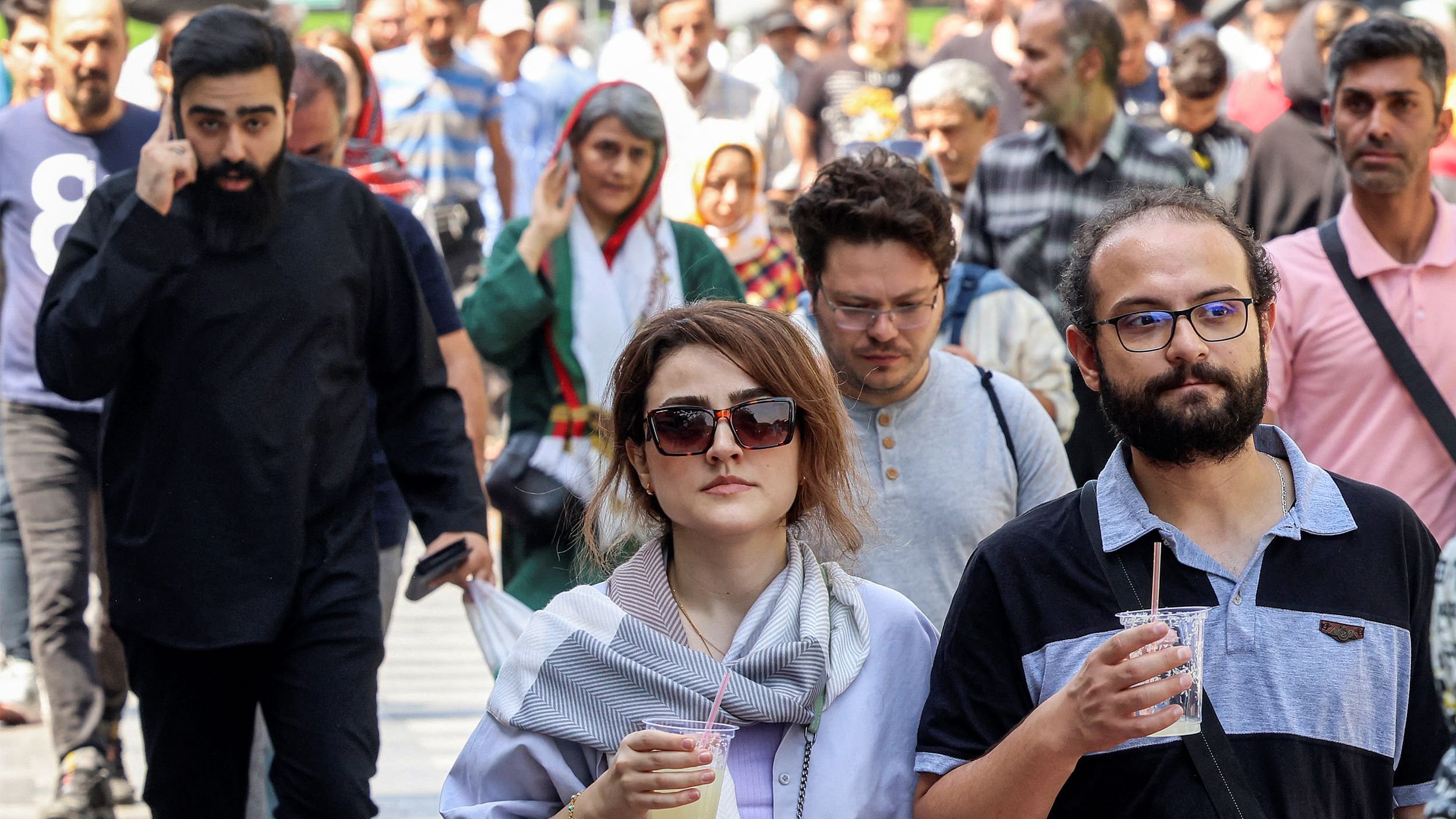 الصورة تظهر مجموعة من الأشخاص يسيرون في الشارع. في المقدمة، هناك امرأتان تحملان مشروبًا، بينما خلفهن يوجد رجال ونساء آخرون في مجموعات صغيرة. يبدو أن هناك تنوعًا في الأزياء بين الأشخاص، مع بعضهم يرتدي الملابس التقليدية والبعض الآخر يرتدي ملابس حديثة. الجو يبدو مفعمًا بالحركة والنشاط، مما يعكس أجواء الحياة اليومية في المدينة.