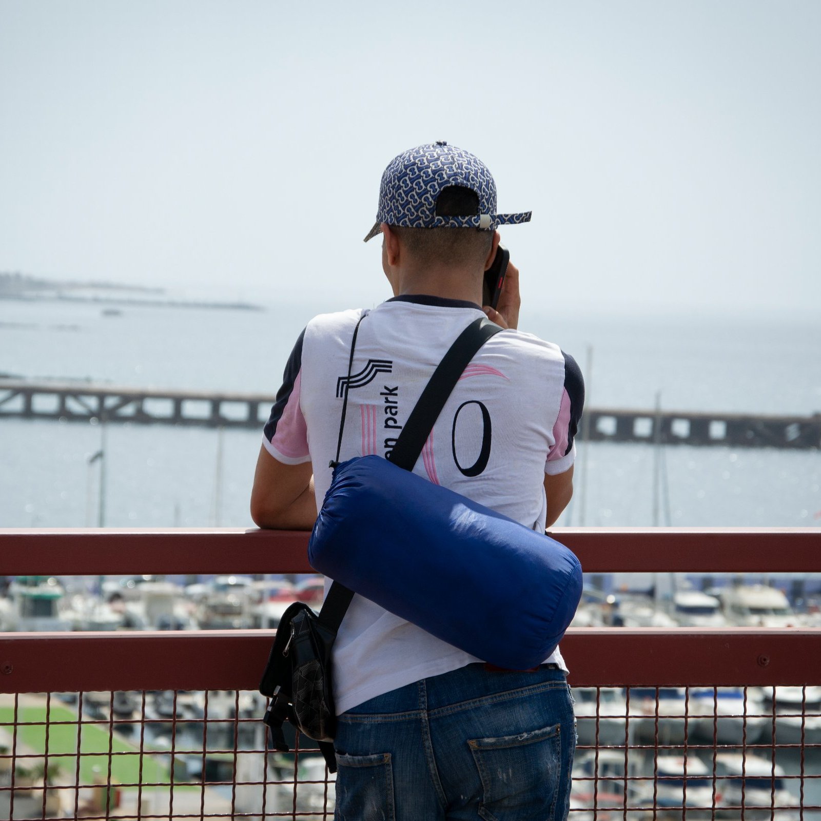 Un homme regarde la mer depuis un pont, avec un port et des bateaux en arrière-plan.