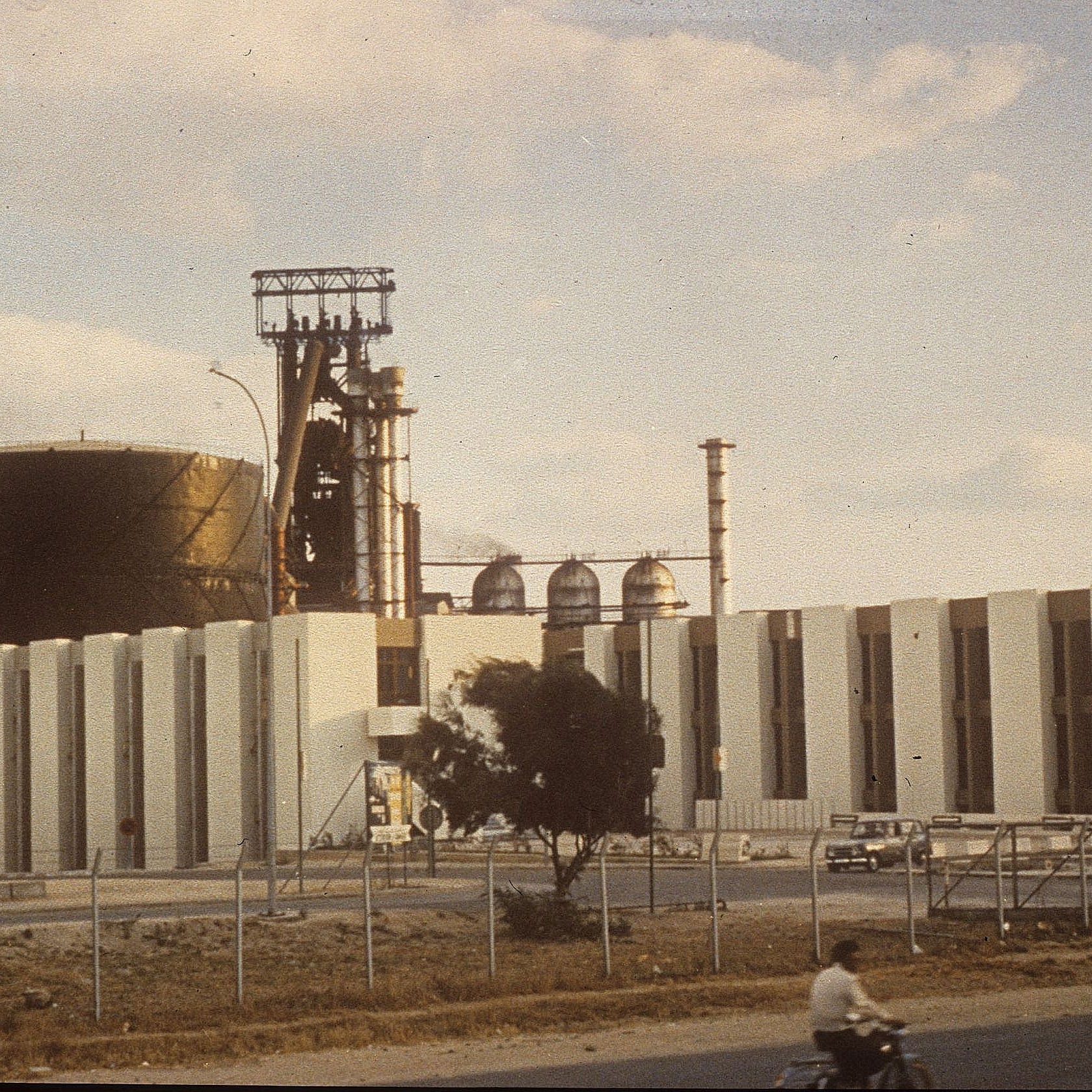L'image montre une grande installation industrielle. On peut voir un grand réservoir noir et une structure avec des cheminées et des tubes. La façade de l'immeuble est faite de blocs blancs. À l'avant, il y a une route, et on aperçoit une personne à vélo sur celle-ci. L'environnement semble être un site industriel, avec quelques arbres et une clôture qui délimite la zone. Le ciel est partiellement nuageux, ce qui donne une ambiance légèrement vintage à la scène.