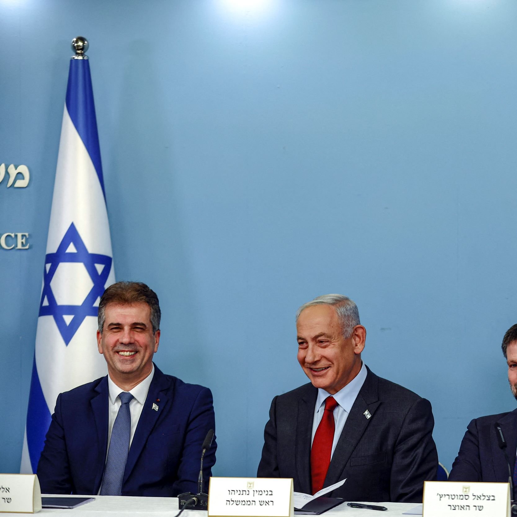 تصویر نشان‌دهنده یک جلسه رسانه‌ای یا کنفرانس خبری است که در آن چهار مرد در یک میز قرار دارند. در پس‌زمینه، پرچم اسرائیل دیده می‌شود. نفر اول از چپ به سمت راست لبخند می‌زند و ظاهراً شاداب است. نفر دوم، که در وسط نشسته، چهره‌ای جدی‌تر دارد و به نظر می‌رسد سخنگوی اصلی جلسه باشد. نفر سوم، که کمی از افراد دیگر فاصله دارد، یک نیمرخ متمرکز دارد و نفر چهارم در انتها نیز به سمت دوربین نگاه می‌کند. فضای تصویر رسمی به نظر می‌رسد و نمایشگر تعامل سیاسی یا اجتماعی است.