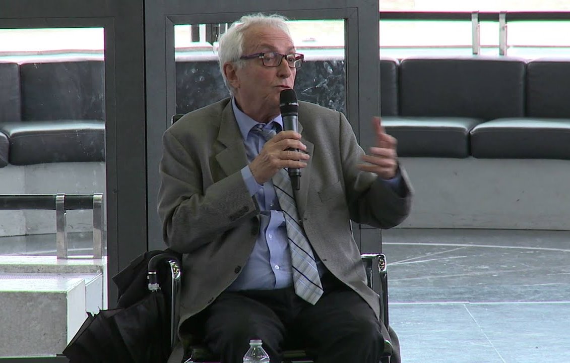 در این تصویر، فردی در حال صحبت کردن با میکروفن است. او کت و شلوار به تن دارد و عینک می‌زند. به نظر می‌رسد در یک جلسه یا کنفرانس شرکت کرده و به حاضران ارائه‌ای می‌دهد. زمینه تصویر نشان‌دهنده یک فضای مدرن و باز است.