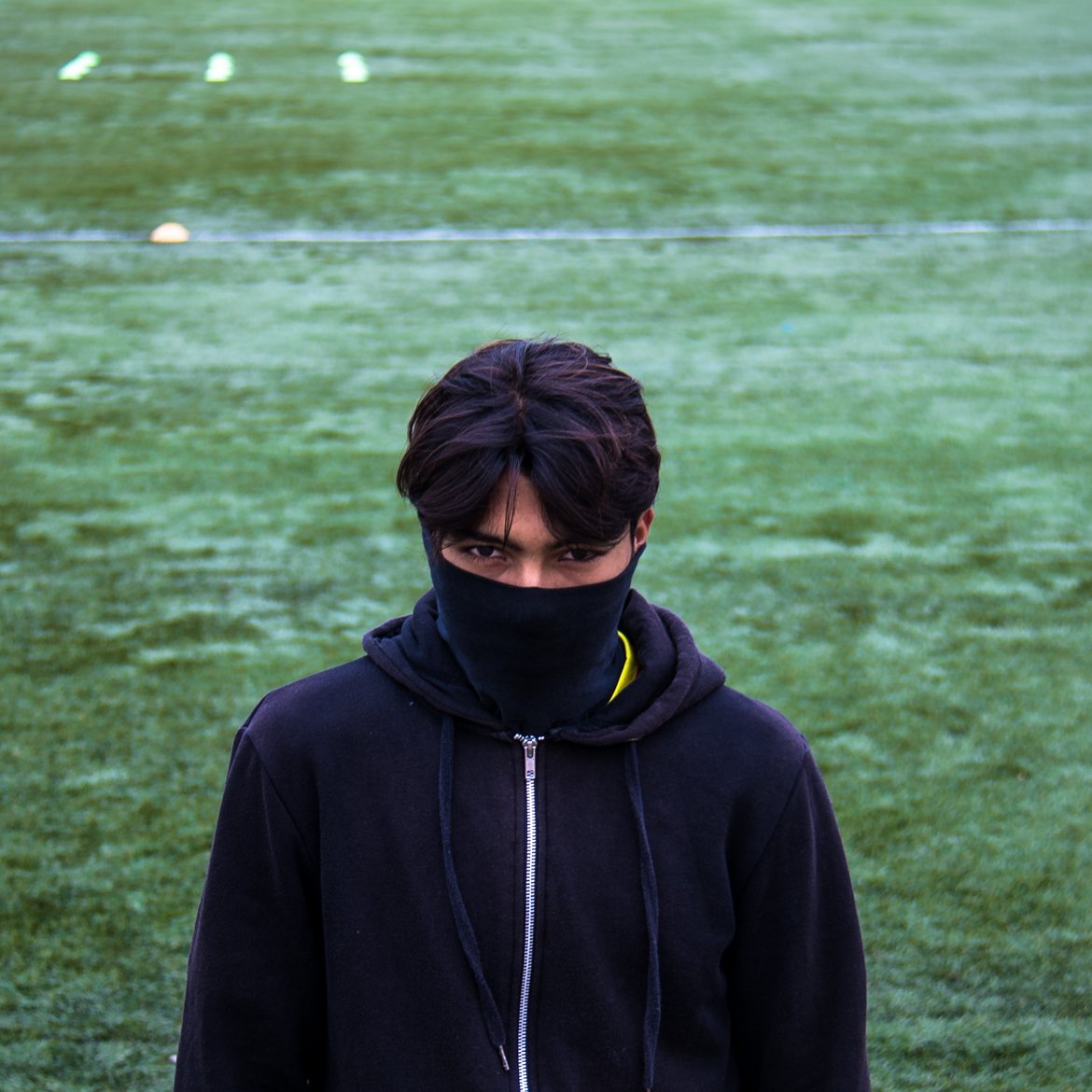 The image features a young person standing on a green sports field, possibly a soccer pitch. They are wearing a black hoodie and have a black mask covering the lower part of their face. The focus is on their intense gaze, and they appear to be looking directly at the camera. The field is marked with white lines and has some yellow cones in the background, suggesting a training or practice session. The overall mood is somewhat mysterious and serious.