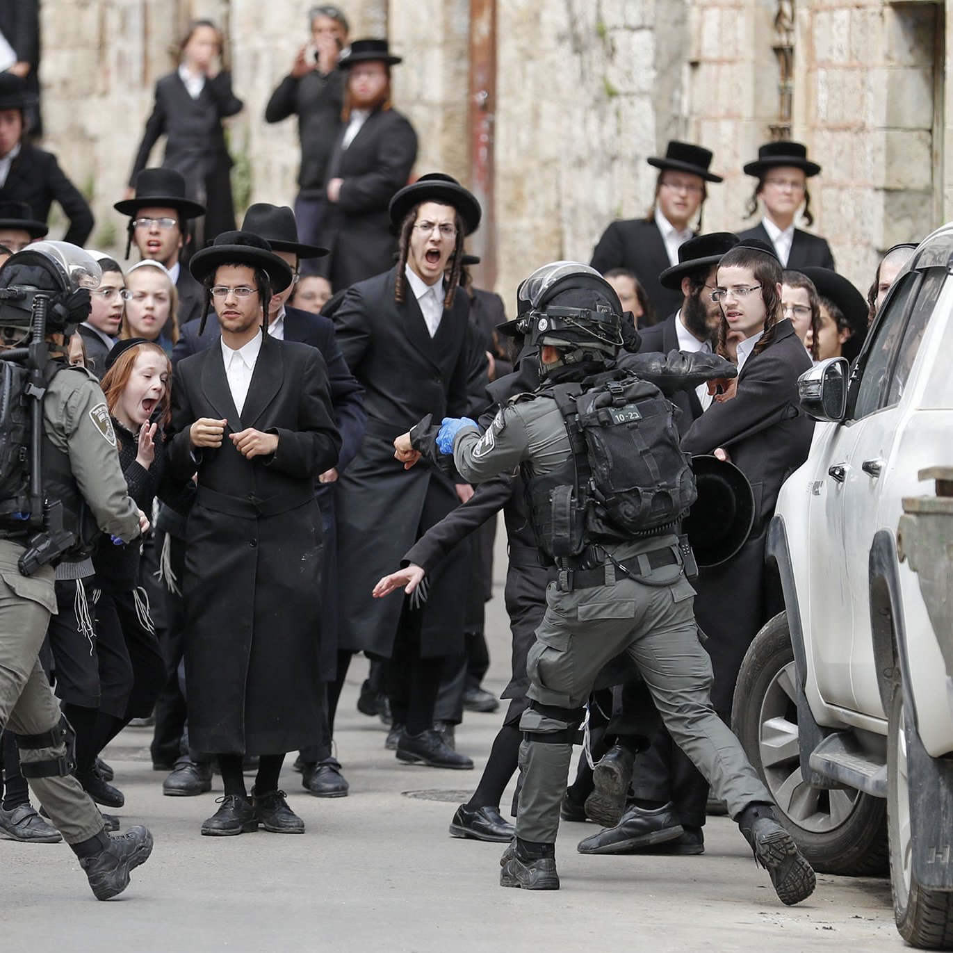 The image depicts a tense confrontation between a group of individuals dressed in traditional Orthodox Jewish attire and armed security personnel. The group appears to be expressing strong emotions, possibly protest or unrest, while the security officers, equipped with tactical gear, seem to be engaging with them. The setting appears to be an urban area, with a mixture of traditional and modern elements visible in the background, including vehicles and stone buildings. The scene captures a moment of conflict or significant social interaction.
