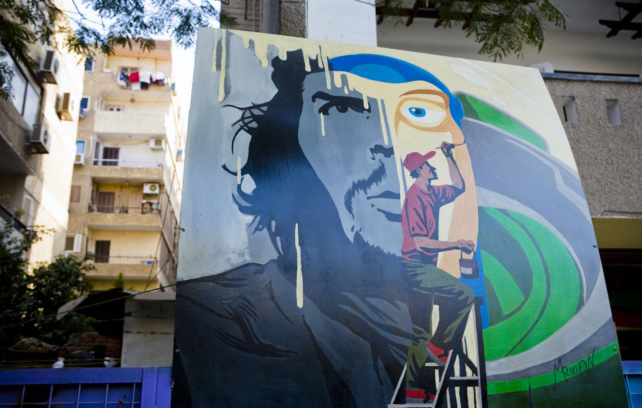 L'image montre une fresque murale colorée. À gauche, il y a le visage d'un homme avec une expression intense, tandis qu'à droite, un artiste est représenté en train de peindre. L'artiste est vêtu d'un pantalon rouge et d'un t-shirt, et il est sur une échelle, concentré sur son œuvre. En arrière-plan, on aperçoit des bâtiments avec des balcons et du linge suspendu. Les couleurs de la fresque sont vives, avec des nuances de bleu et de vert qui contrastent avec les éléments plus sombres du visage.