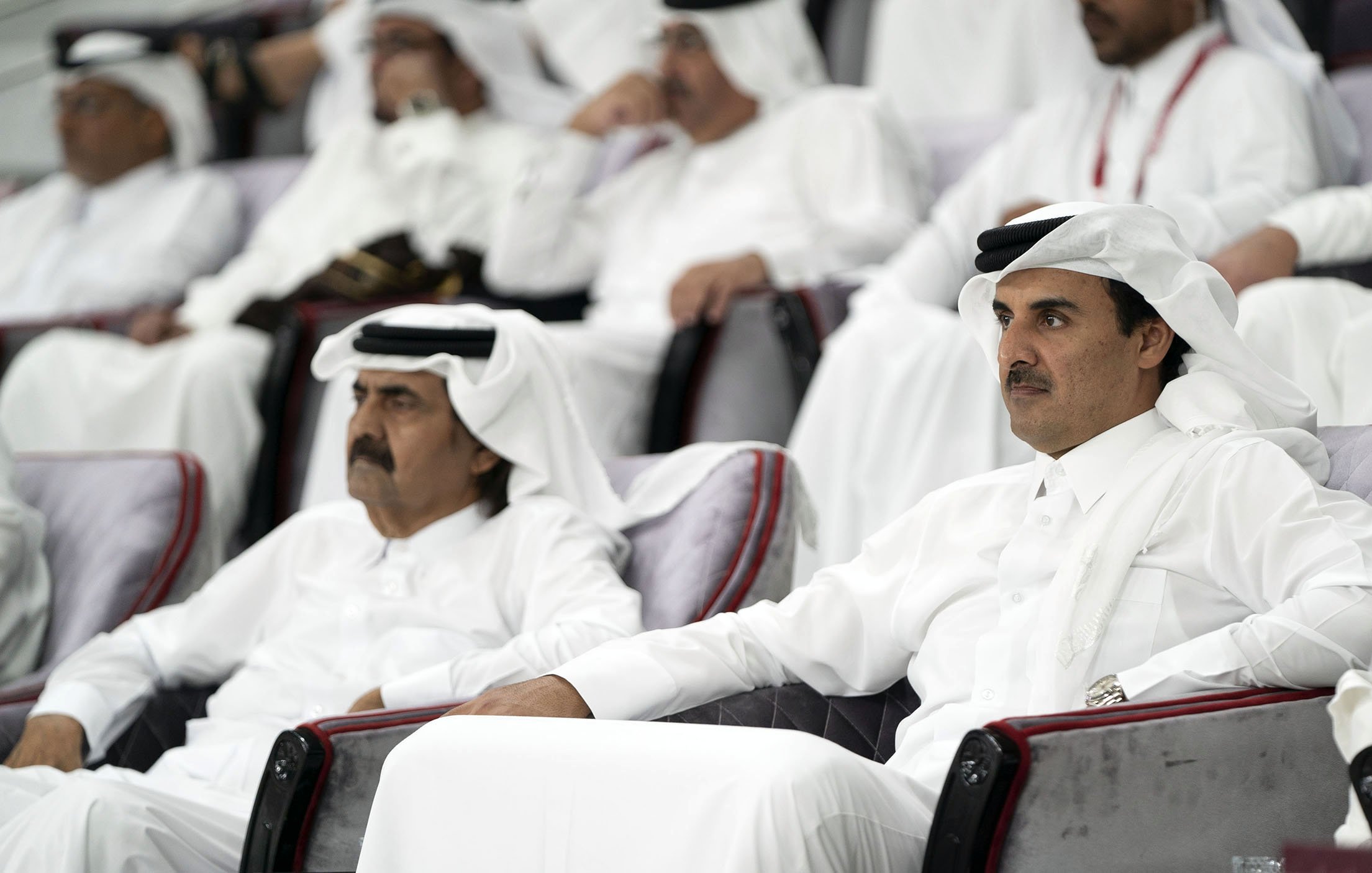 در این تصویر، دو مرد در حالی که لباس‌های سنتی عربی، شامل قمیص و عقال، پوشیده‌اند، نشسته‌اند. آن‌ها در یک مکان رسمی به تماشای چیزی مشغول‌اند. پس‌زمینه افراد دیگری را نشان می‌دهد که همچنین در لباس‌های سنتی نشسته‌اند. فضای کلی تصویر جدی و متمرکز به نظر می‌رسد.