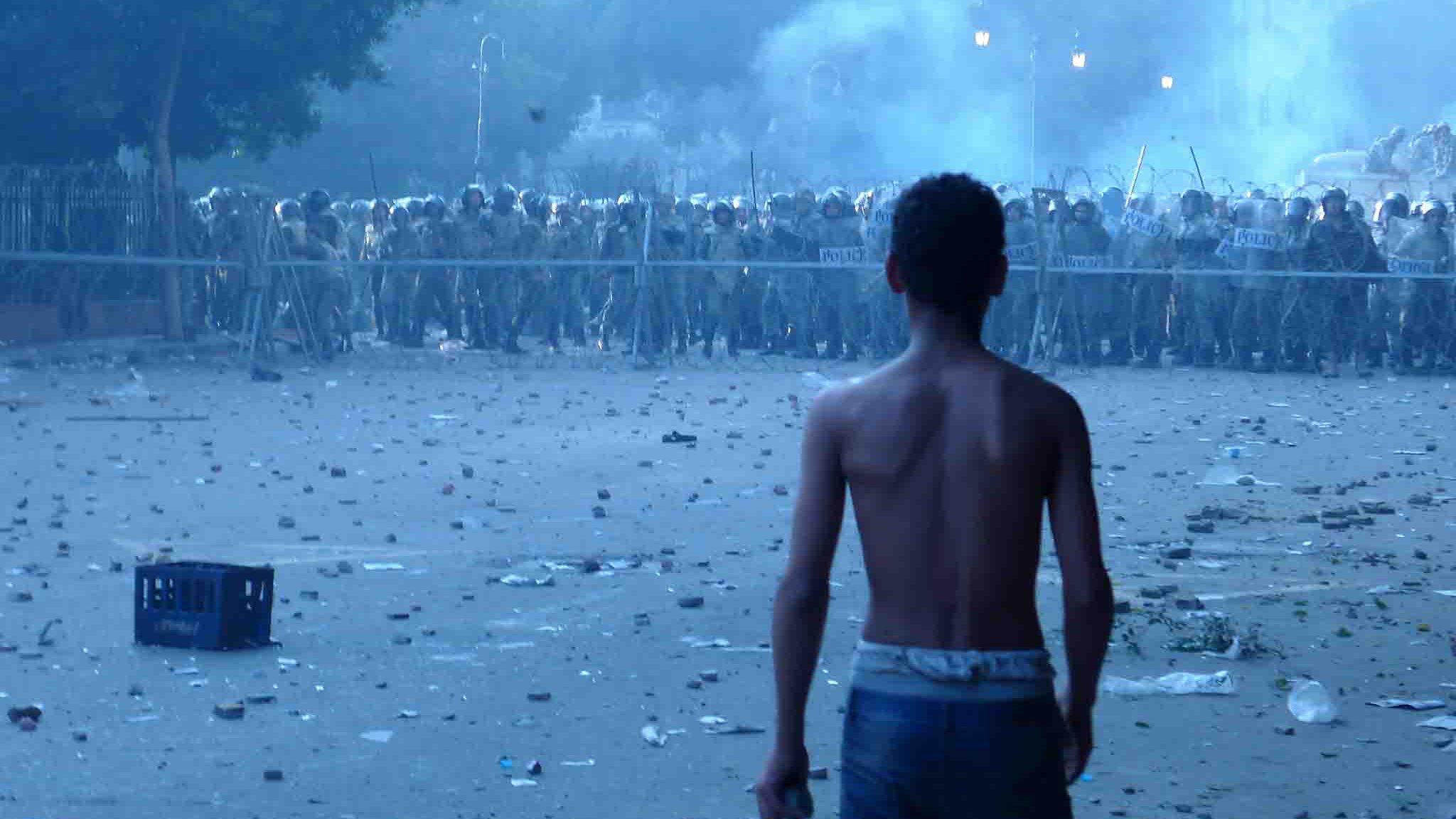 L'image montre un jeune garçon, torse nu, qui se tient de dos face à une ligne de forces de l'ordre en uniforme. Il est dans un environnement troublé, avec des débris et des bouteilles éparpillés au sol, ainsi qu'une légère brume qui donne une atmosphère tendue et chaotique. La scène semble se dérouler dans un contexte de troubles ou de manifestations. Les policiers, en arrière-plan, se tiennent en formation, suggérant un affrontement imminent.