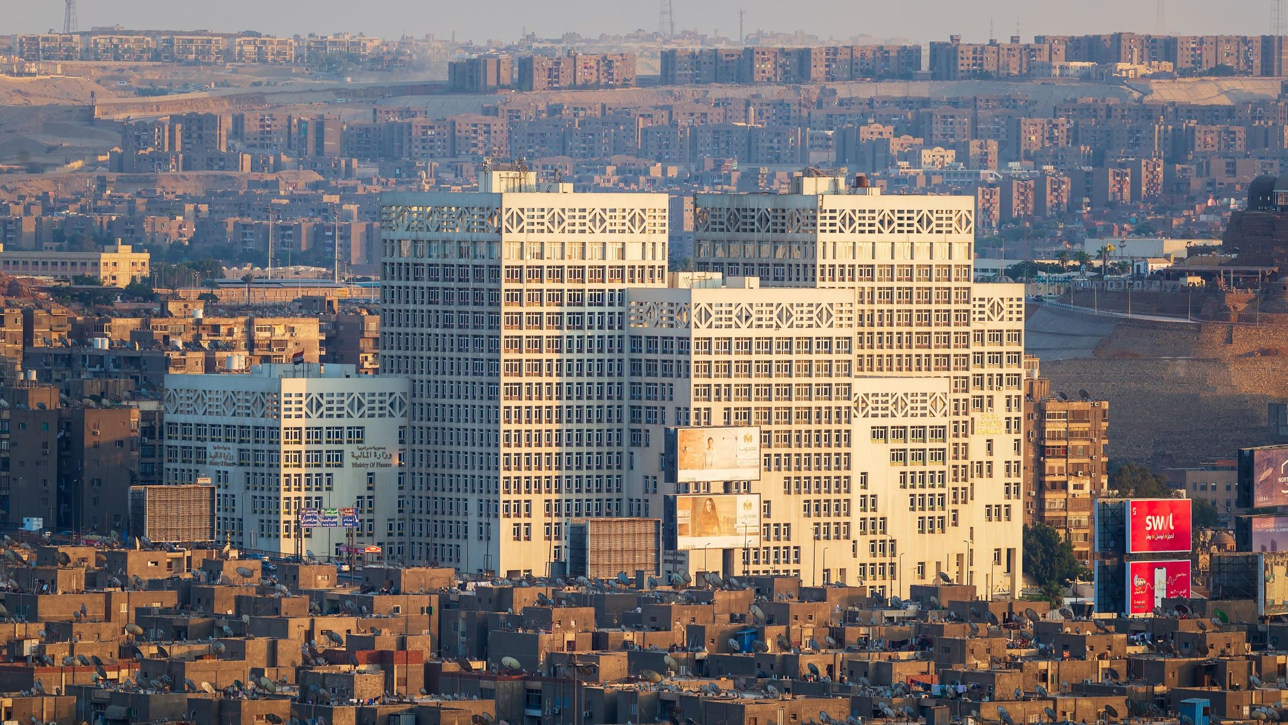 La imagen muestra una vista panorámica de una ciudad con edificios modernos en primer plano. Los edificios tienen un diseño arquitectónico distintivo y están iluminados por la luz del sol, lo que resalta su estructura. Al fondo, se pueden ver más edificios y una serie de montañas o colinas, lo que sugiere un paisaje urbano extenso. La atmósfera es clara, y la escena transmite una sensación de desarrollo urbano y modernidad.