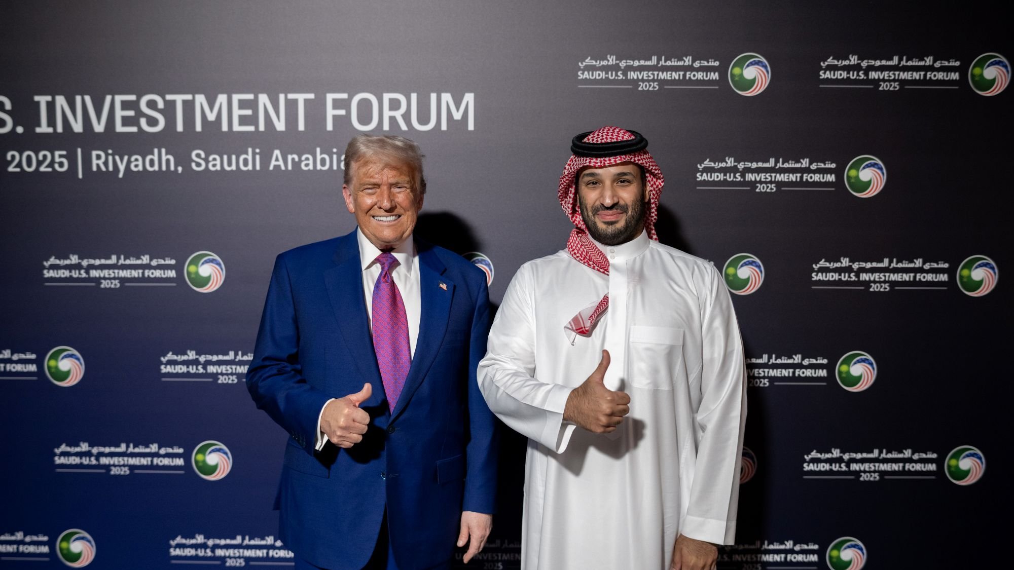 دو مرد در لباس رسمی، یکی با کت و شلوار و دیگری با لباس سنتی عربی، لبخند می‌زنند و اشاره به عالی بودن دارند.